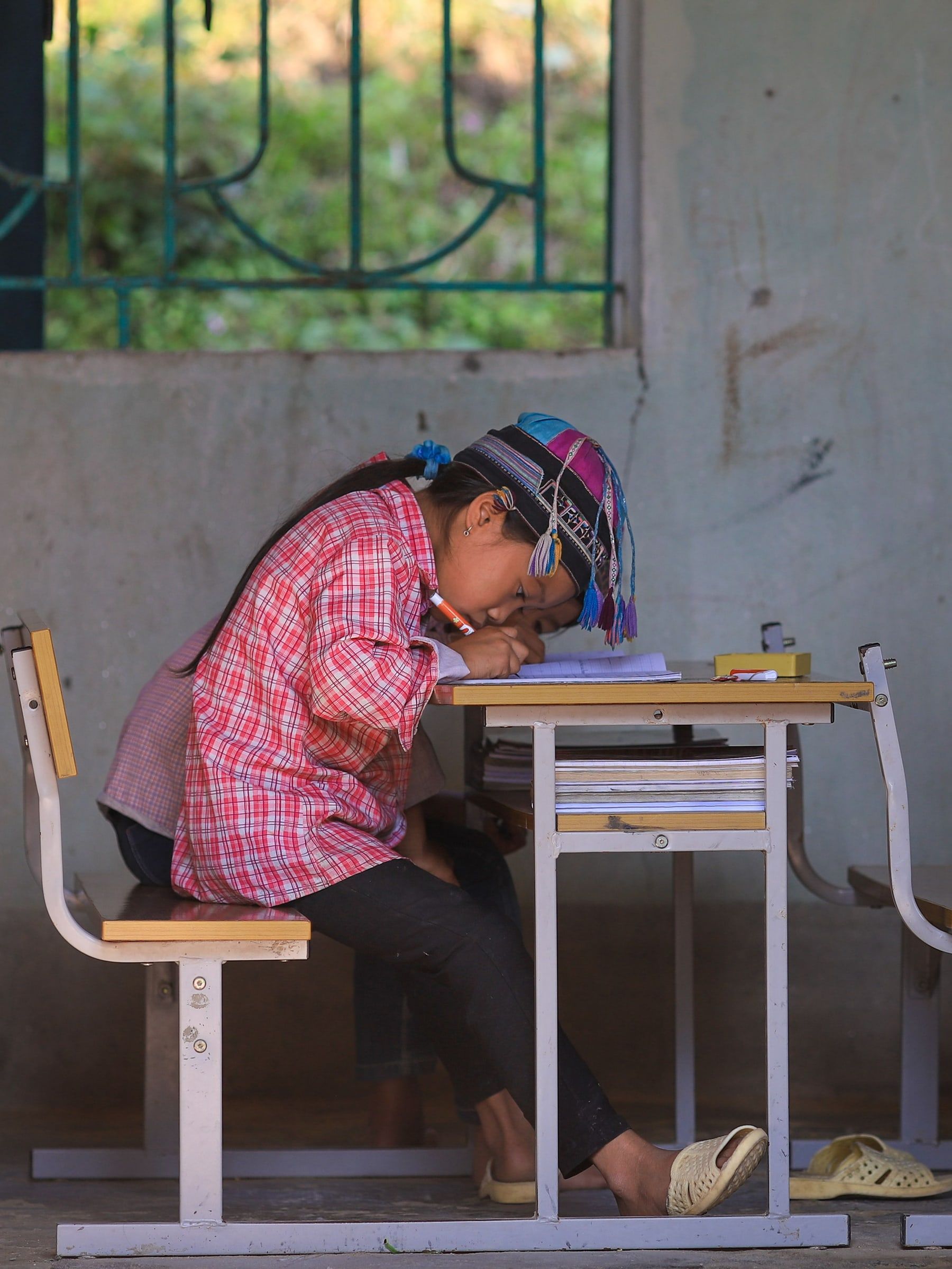 Một em nhỏ mặc áo kẻ đỏ, đội mũ thổ cẩm, cúi viết bài bên bàn học trong lớp.