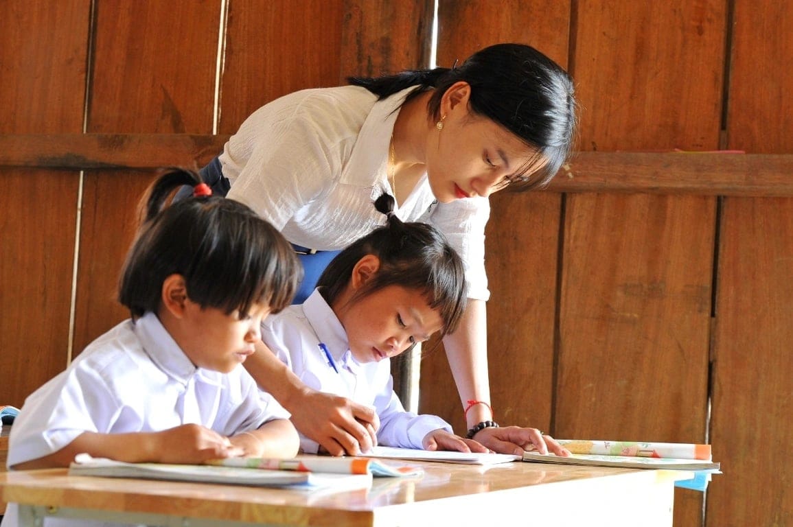 Một cô giáo cúi xuống hướng dẫn hai em nhỏ viết bài.